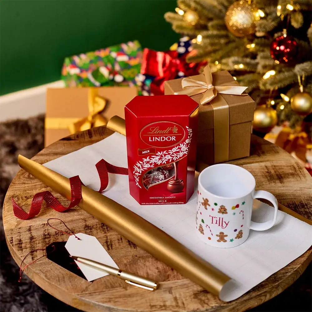 Christmas gift wrapping scene with chocolates, a personalised festive mug, wrapped presents, gift tags, and wrapping paper beside a decorated Christmas tree. Christmas gift wrapping scene with chocolates, a personalised festive mug, wrapped presents, gift tags, and wrapping paper beside a decorated Christmas tree.